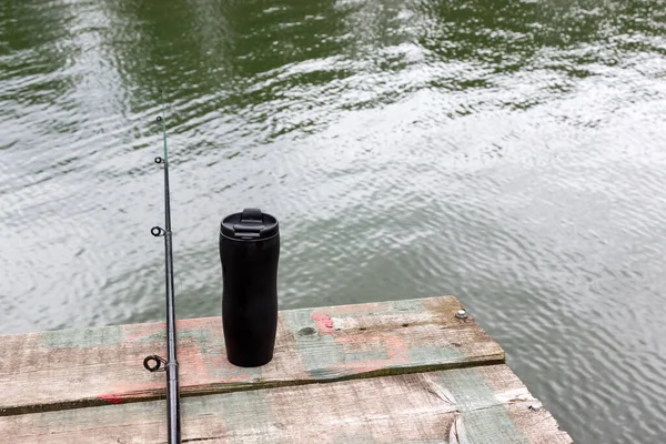fishing rod is lying on a wooden pier hanging over the water and there is a black thermos of tea nearby. concept for the day of the fisherman. Outdoor recreation and food on nature
