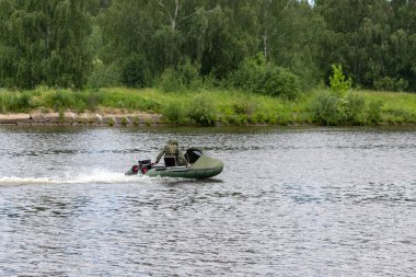 Şişme motorlu teknedeki balıkçı nehirde yüzüyor. Yeşil sahilde, köy evleriyle birlikte nehirde yüzen bir teknede kamuflajlı bir adam. Gölde sonbahar balıkçılığı. Eğlence amaçlı balıkçılık yasağı kavramı