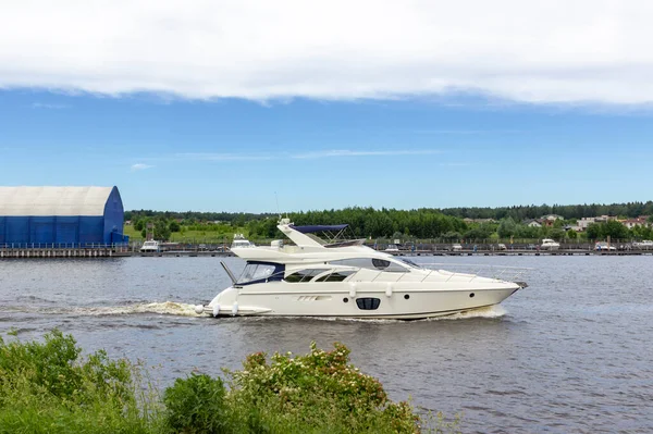 a long luxurious yacht sails along the riverbed against the background of the coastline and the hangar for boats. The concept of Day of the Navy and fishing with friends.Sunny weather and blue sky.