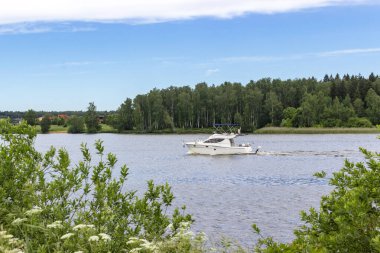 Bir motorlu tekne mavi bir gökyüzü ve güneş ışığı altında nehir boyunca yol alır. Yeşil bir orman ve kırsal bir yerleşim yerinin yakınında. Göz kamaştırıcı bir manzara ve iyi bir çekim açısı..