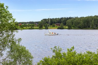 Mavi gökyüzü ve güneş ışığı altında nehir boyunca yelken açan bir yat. Yeşil bir orman ve kırsal bir yerleşim yerinin yakınında. Göz kamaştırıcı bir manzara ve iyi bir çekim açısı..