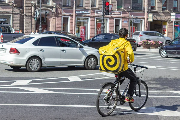 Rusya, Saint Petersburg, 09.08.2020. Yandex yemek kuryesi kırmızı ışıkta karşıya geçiyor, trafik kurallarını ihlal ediyor. Bisikletli sarı sırt çantalı teslimatçı. Teslimat şirketi.