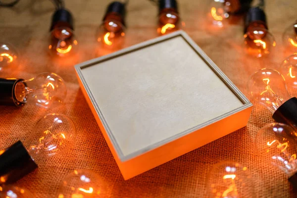 Box of plywood on the background of burlap. Around the box is a retro garland with Edison lamps. Free space.