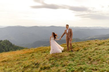 Gelin ve damat birbirlerine nazikçe sarılıyorlar. Gün batımı. Sonbahar dağlarının arka planında düğün fotoğrafı. Güçlü bir rüzgar saç ve elbise şişirir. Yakın plan.