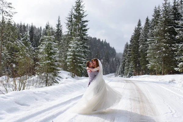 The groom circles the bride in his arms on the background of a snowy fir forest. Snowing. Winter wedding.