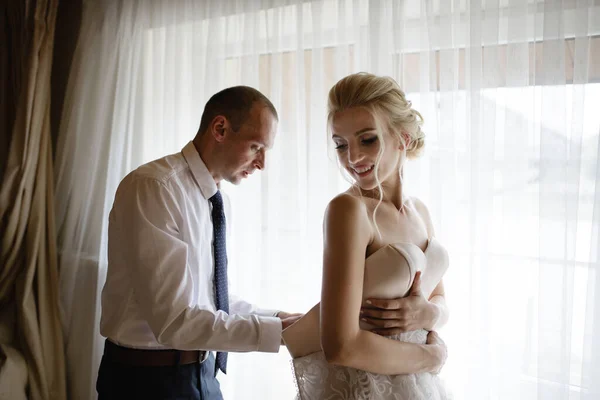 漂亮的金发碧眼的新娘穿着白色华丽的婚纱 准备参加婚礼 早上的准备工作一个女人穿上一件衣服 新郎帮助新娘 — 图库照片