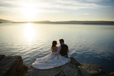 Gelin ve damat bir uçurumun kenarında gölün arka planında oturuyorlar..