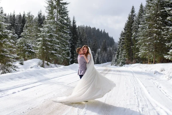 The groom holds in his arms and circles his bride. Winter wedding. A couple stands in the background of a snowy forest.