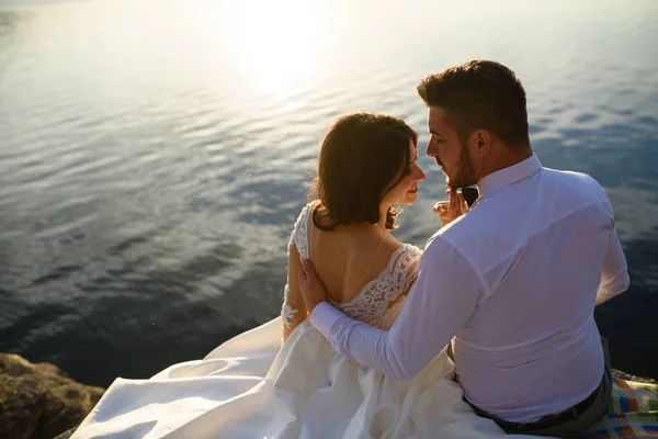 新娘和新郎正靠着湖的背景坐在悬崖边上 — 图库照片