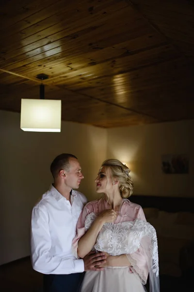 The bride in a silk bathrobe is trying on her dress. She is hugged by her fiance. The bride and groom are preparing for the wedding together.