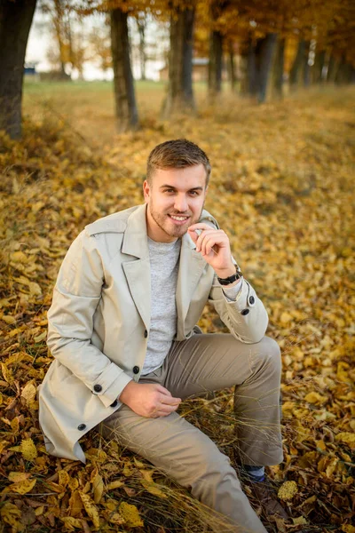 一个穿着外套 背景在秋天树上的年轻人的画像 一个男人坐在地上 — 图库照片