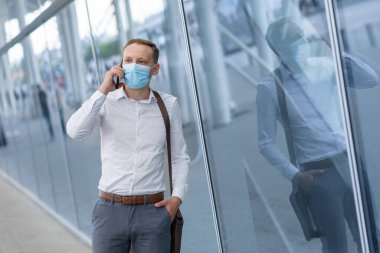 Genç bir işadamı, modern bir ofis binasının arka planında tıbbi bir maskeyle telefonda konuşuyor..