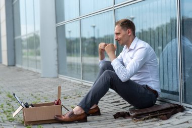 Kovulmuş bir ofis çalışanı modern bir ofis binasının yanında yerde oturuyor. Adam işten çıkarılma konusunda çok endişeli. Çalışan çok kızgın..