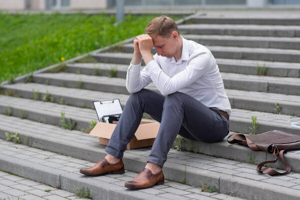A fired office worker sits on the steps. The man does not know what to do next. Next to it is a cardboard box with stationery.
