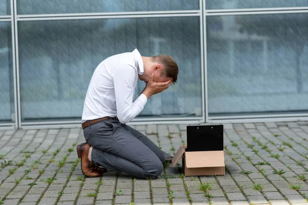 The fired office worker fell to his knees and covered his face due to stress. In front of him is a cardboard box with stationery. The man is unhappy due to a reduction in the robot.