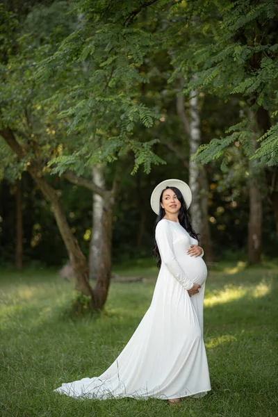 緑の木の背景に白いドレスを着た帽子をかぶった妊婦 — ストック写真