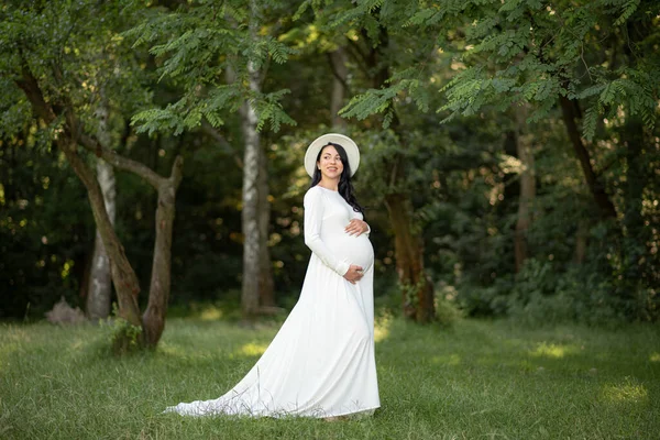 Беременная Женщина Шляпе Позирует Белом Платье Фоне Зеленых Деревьев — стоковое фото