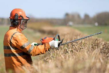 Çit Kesme Hizmetleri. Profesyonel Bahçıvan Üniformalı ve İşitme Koruyucu Kulaklıklı İşçi Hedgerow 'u Gas Power Clipper ile buduyor.
