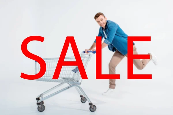 Glücklicher junger Mann läuft mit Einkaufswagen und lächelt in die Kamera auf grau, Verkaufsaufschrift — Stockfoto