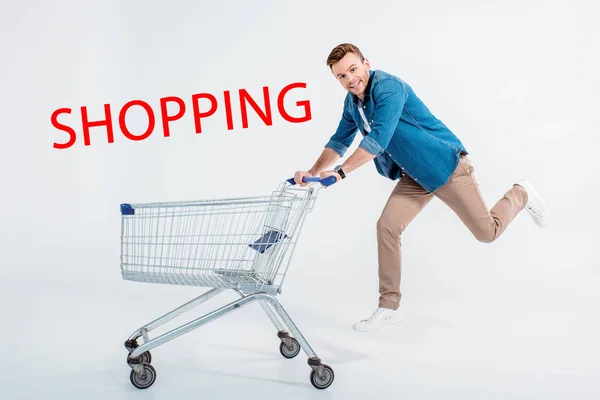 Joven feliz corriendo con carrito de compras y sonriendo a la cámara en gris, inscripción de compras - foto de stock