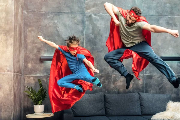 Pai e filho em capas de super-heróis e máscaras pulando no sofá em casa — Fotografia de Stock