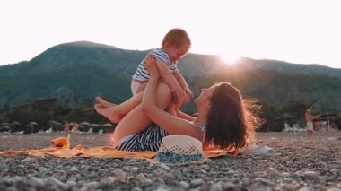 Genç anne küçük bebek kızı ile Dağların üzerinden yaz gün batımında sahilde oturan eğlenin. Mutlu aile kavramı. Mutlu çocukluk. HD video görüntüleri ağır çekimde tam