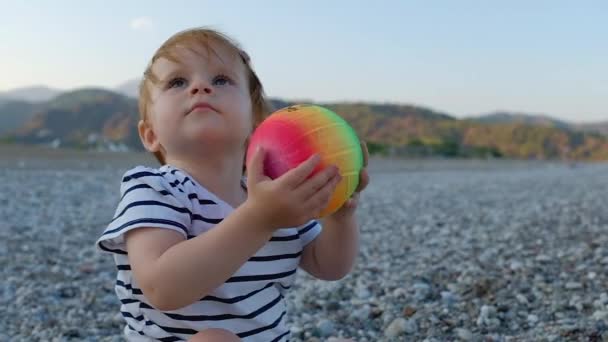 niedliches kleines Mädchen mit buntem Ball in den Händen, das an einem Sommertag am Strand sitzt. Glückliche Kindheit. Full-HD-Zeitlupenaufnahmen. 1920x1080 Dolly-Schuss