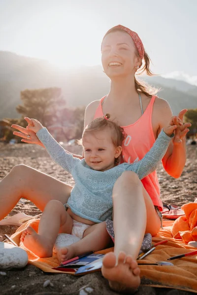 Молода мати зі своєю маленькою дівчинкою весело сидить на пляжі в літній день на заході сонця над горами. Щаслива сімейна концепція. Щасливе дитинство — стокове фото