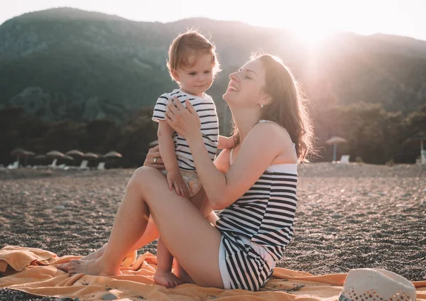 Молодая мать со своей маленькой девочкой весело провести время, сидя на пляже в летний день на закате над горами. Счастливая семейная концепция. Счастливого детства — стоковое фото