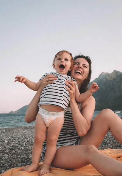 La madre joven con su pequeña niña se divierte sentada en una playa en el día de verano al atardecer sobre las montañas. Concepto familiar feliz. Feliz infancia. —  Fotos de Stock
