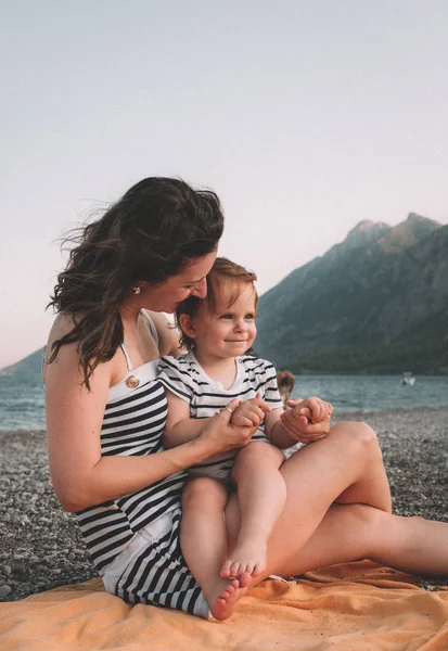 Молода мати зі своєю маленькою дівчинкою весело сидить на пляжі в літній день на заході сонця над горами. Щаслива сімейна концепція. Щасливе дитинство — стокове фото
