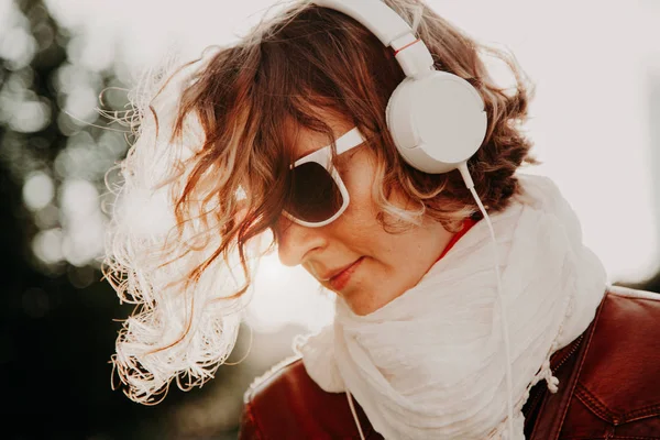 Красива молода жінка в сонцезахисних окулярах і навушниках слухає музику проти яскравого заходу сонця. Концепція випадкового стилю — стокове фото