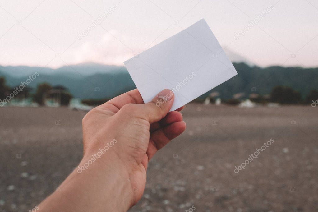 Empty paper note in hand at amazing landscape background. Travel and Vacation concept. White piece of paper with no text.