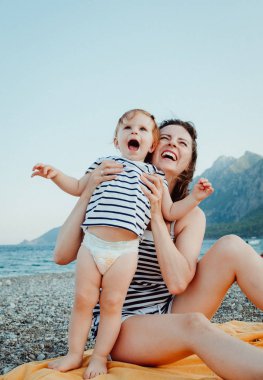Genç anne küçük bebek kızı ile Dağların üzerinden yaz gün batımında sahilde oturan eğlenin. Mutlu aile kavramı. Mutlu çocukluk