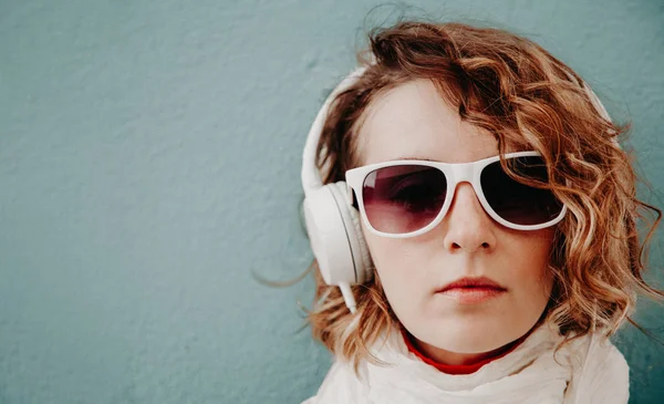 Hermosa joven con gafas de sol y auriculares escuchando música de pie en la pared. Concepto de estilo casual — Foto de Stock