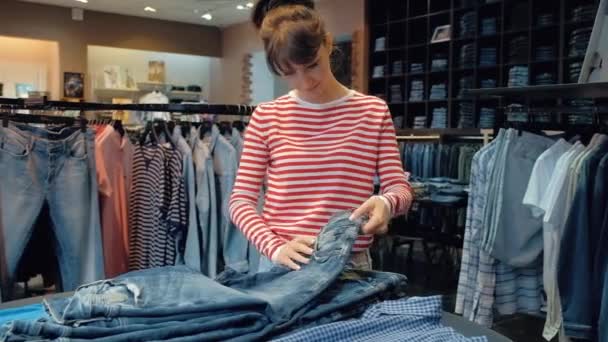 Молодая продавщица в джинсовой лавке перелистывает стопку джинсовых штанов — стоковое видео