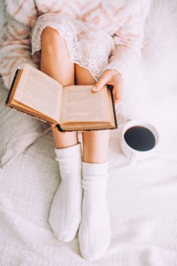 Genç bir kadın, eski bir kitap ve bir fincan kahveyle yatakta kalmaktan zevk alıyor..