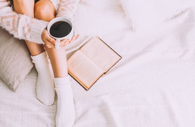 Genç bir kadın, eski bir kitap ve bir fincan kahveyle yatakta kalmaktan zevk alıyor..