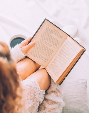 Genç bir kadın, eski bir kitap ve bir fincan kahveyle yatakta kalmaktan zevk alıyor..