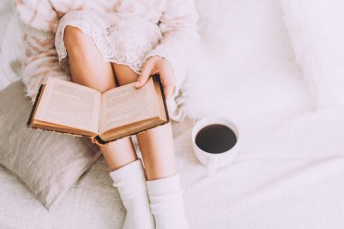 Genç bir kadın, eski bir kitap ve bir fincan kahveyle yatakta kalmaktan zevk alıyor..