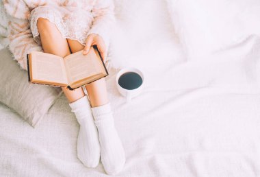Genç bir kadın, eski bir kitap ve bir fincan kahveyle yatakta kalmaktan zevk alıyor..