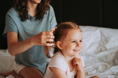 Çekici küçük kız kameraya bakıyor ve gülümsüyor güzel genç annesi kızının saçını tararken.