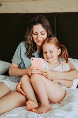 Anne ve kızı yatakta otururken akıllı telefon kullanıyorlar. Mutlu çocuk ve anne iyi eğlenceler..