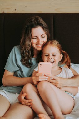 Anne ve kızı yatakta otururken akıllı telefon kullanıyorlar. Mutlu çocuk ve anne iyi eğlenceler..