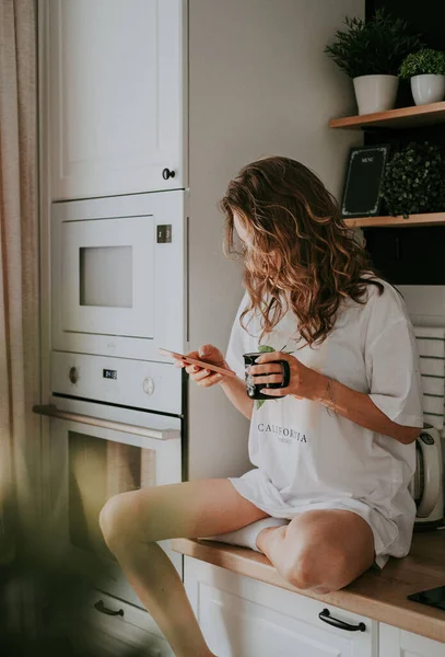 Młoda Ładna Kobieta Pije Kawę Siedząc Kuchni Domu Korzystając Smartfona — Zdjęcie stockowe