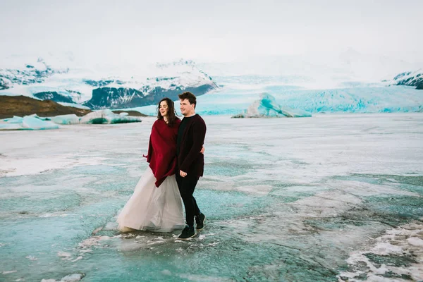 Šťastní novomanželé chůze v ve studené větrný den ledovcové laguny. Islandská svatba. — Stock fotografie