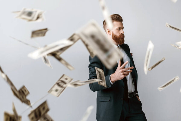 handsome elegant businessman with falling dollar banknotes on foreground isolated on grey