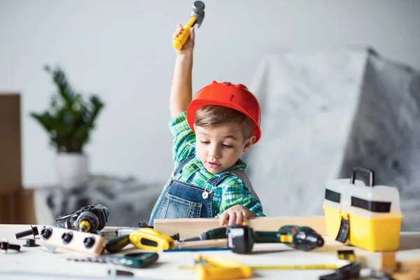 Adorable little kid in red hard hat playing with wooden plank and toy hammer — Stock Photo