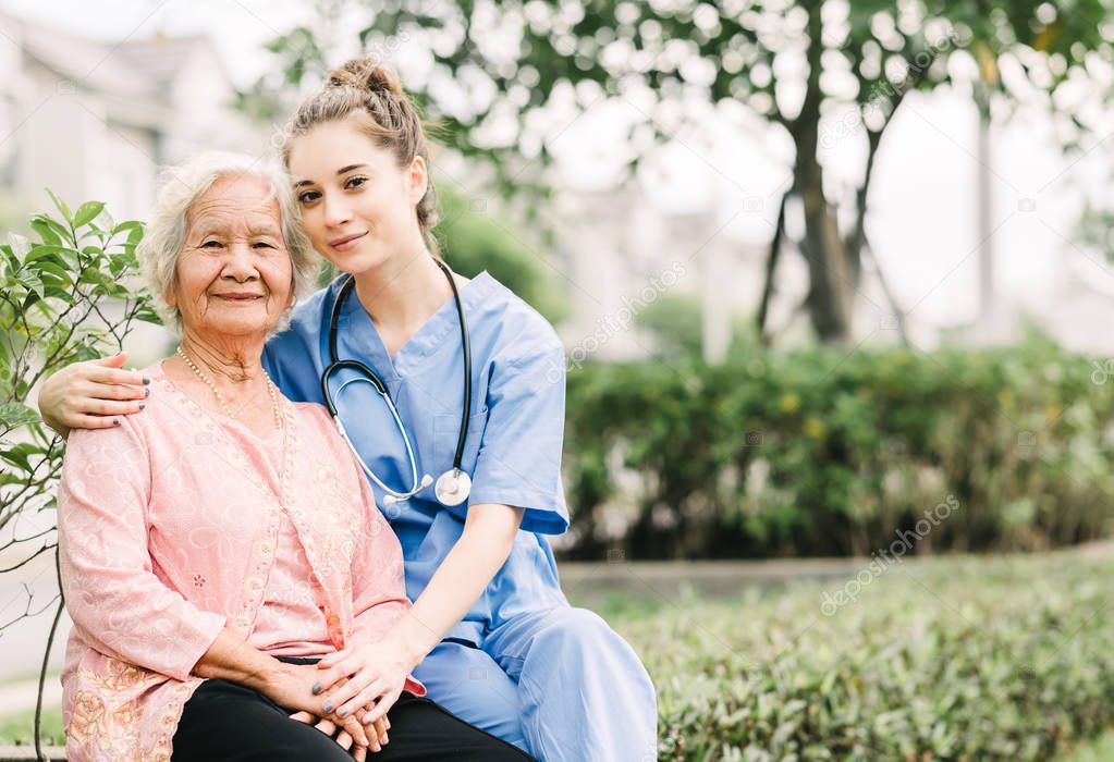 Smiling nurse caregiver embracing happy Asian elderly woman outdoor in the park