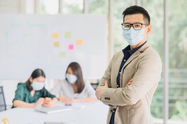 Mutlu Asyalı girişimci yüz maskesi takar. Kollarını kavuşturur ve arka plandaki bir meslektaşıyla modern ofisin toplantı salonunda buluşur. 